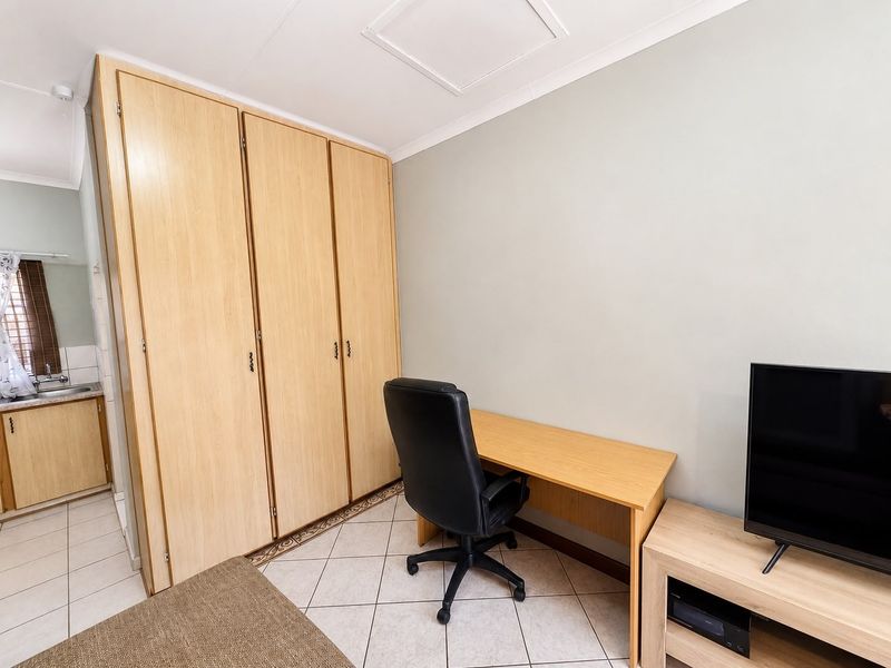 A bedroom featuring built-in wooden cupboards, tiled flooring, and a neutral wall finish, with an adjacent view into a small kitchenette area.