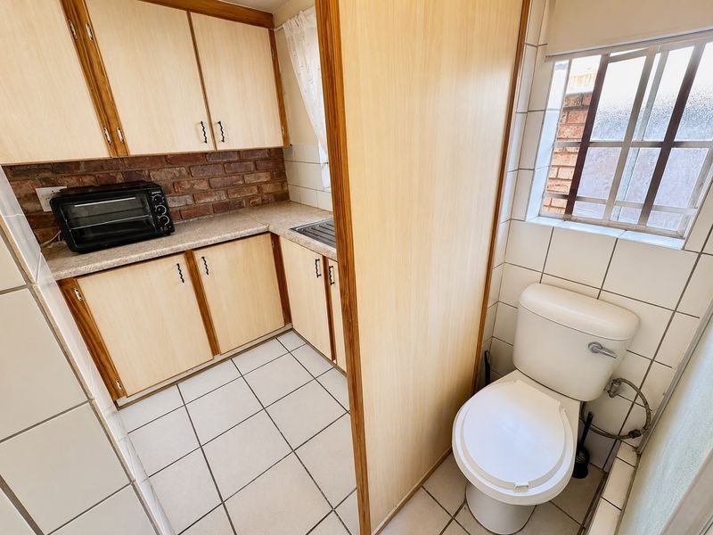 A compact kitchen area featuring laminate cabinetry, brick backsplash, and ceramic tiling, with an adjacent toilet facility.