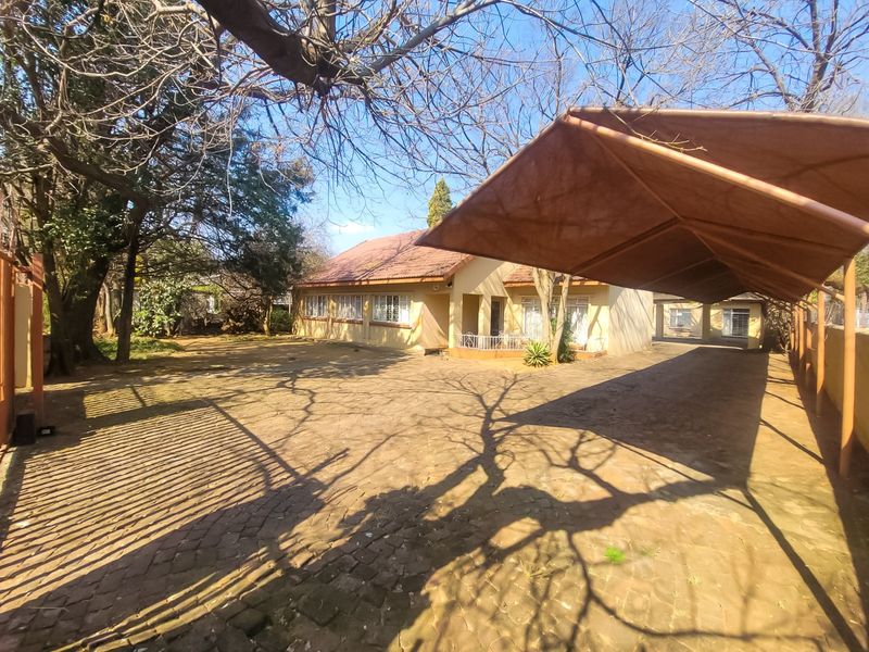 Front exterior view of the house featuring a large paved driveway, a covered carport, and mature trees in the front garden area.