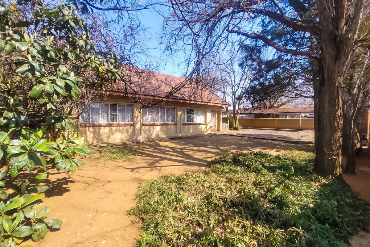 Rear view of the house featuring a tiled roof, brick facade, and a large paved area surrounded by mature trees and garden space.