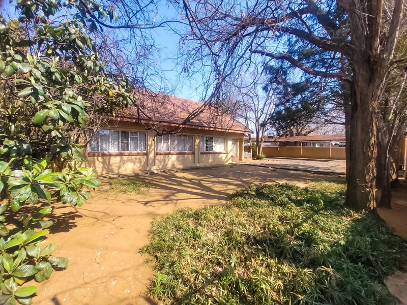 Rear view of the house featuring a tiled roof, brick facade, and a large paved area surrounded by mature trees and garden space.