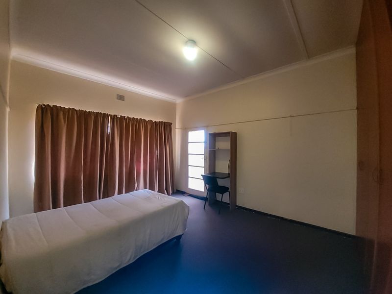 A functional bedroom featuring built-in cupboards, neutral walls, and a large window covered by curtains, providing a simple and practical living space.