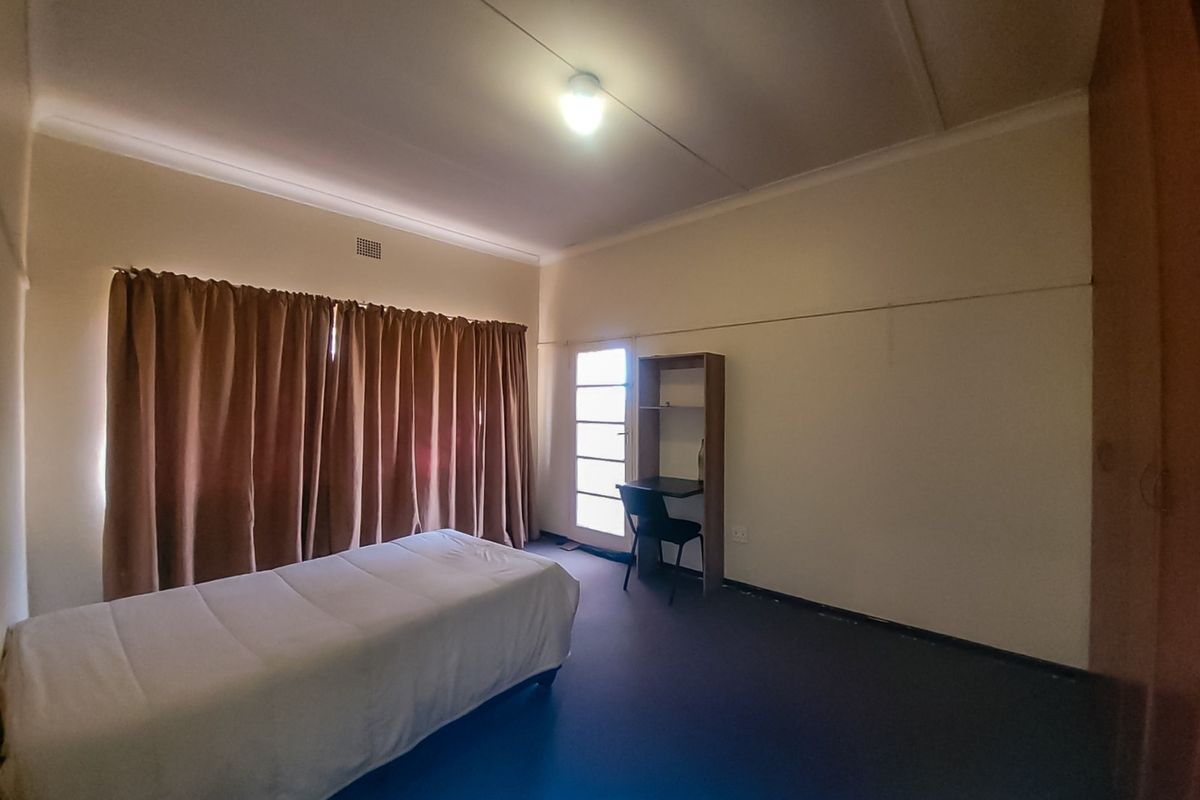 A functional bedroom featuring built-in cupboards, neutral walls, and a large window covered by curtains, providing a simple and practical living space.