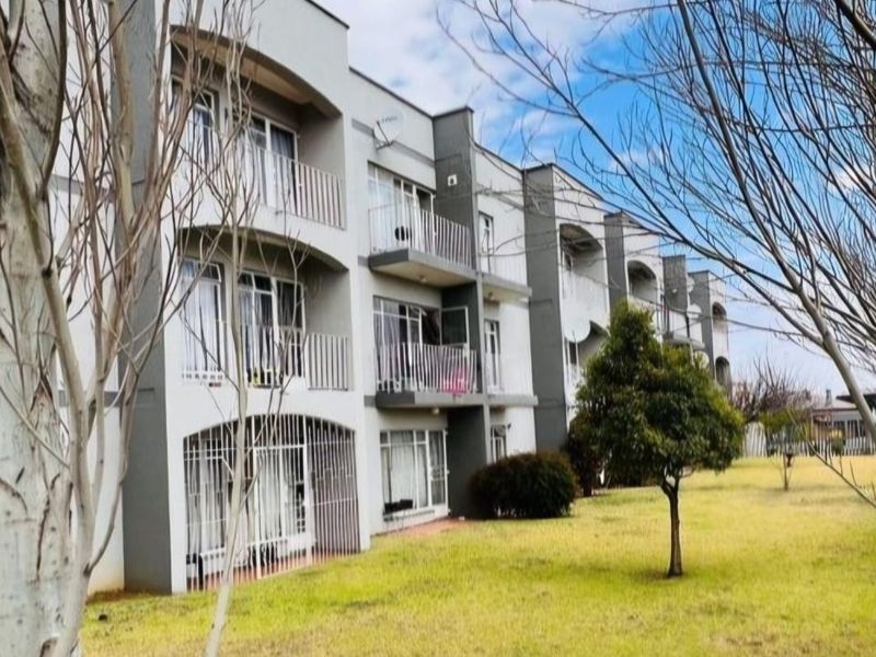 Rear view of a multi-story apartment building featuring private balconies, sliding glass doors, and security bars, set against a landscaped lawn area.
