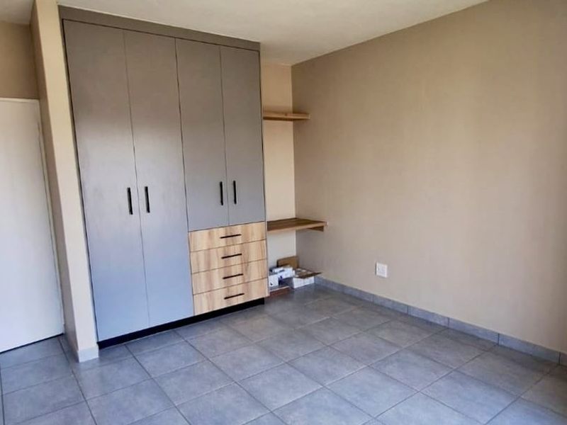 A clean, empty bedroom featuring modern grey and wood-tone built-in cupboards, a matching built-in desk, and neutral tiled flooring.