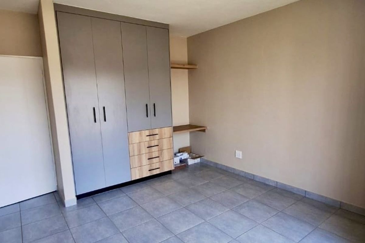 A clean, empty bedroom featuring modern grey and wood-tone built-in cupboards, a matching built-in desk, and neutral tiled flooring.