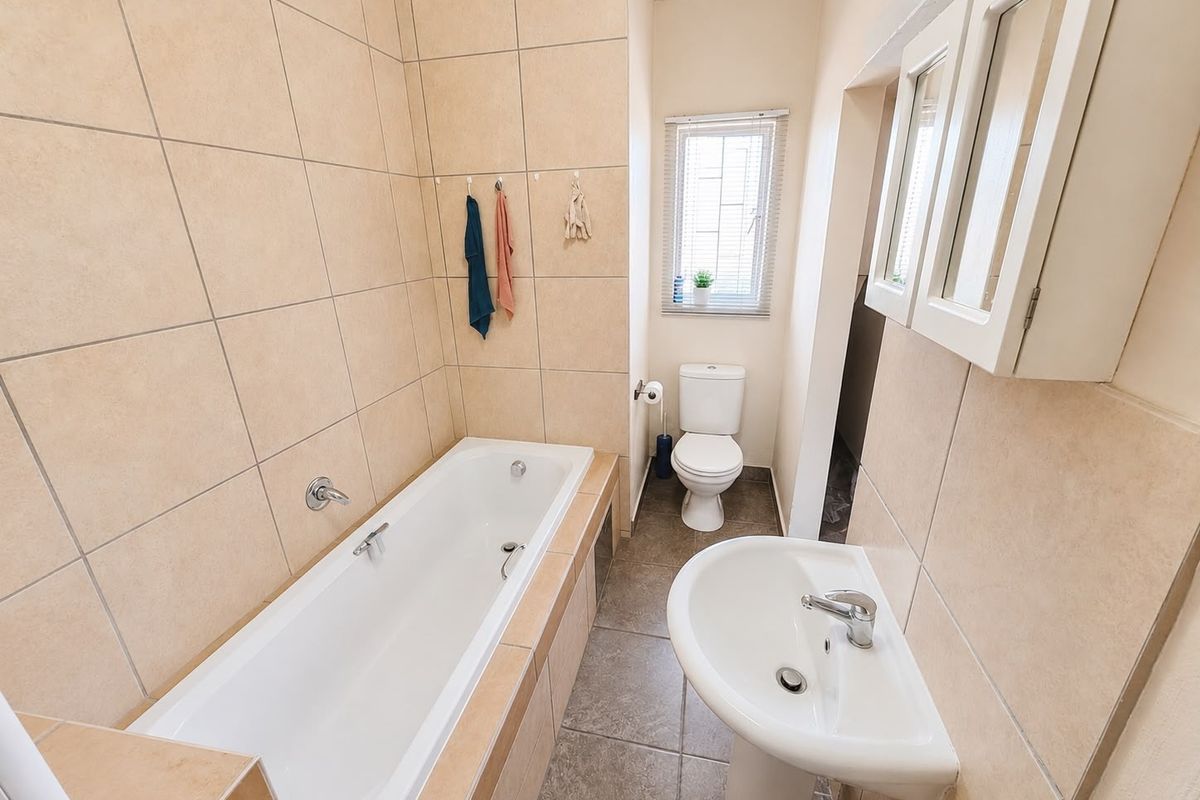 A functional bathroom featuring a tiled bathtub, pedestal basin, toilet, and wall-mounted medicine cabinet with neutral tiling throughout.