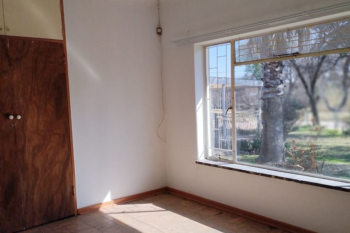 An empty bedroom featuring a built-in wooden wardrobe, tiled flooring, and a large metal-framed window overlooking the garden.
