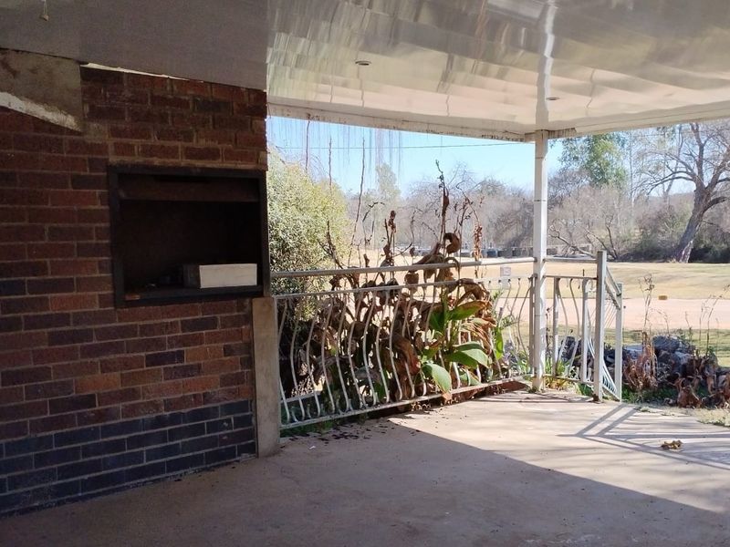 Covered patio area featuring a built-in braai, metal balustrade, and views of the surrounding garden and landscape.