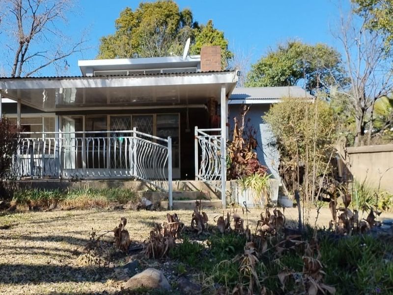 Rear view of the house featuring a covered patio area, metal railings, and a large garden space requiring maintenance.