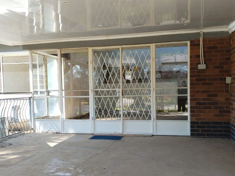 A covered patio area featuring a security gate, sliding glass doors, and brick wall detailing, requiring some maintenance to the flooring and ceiling.