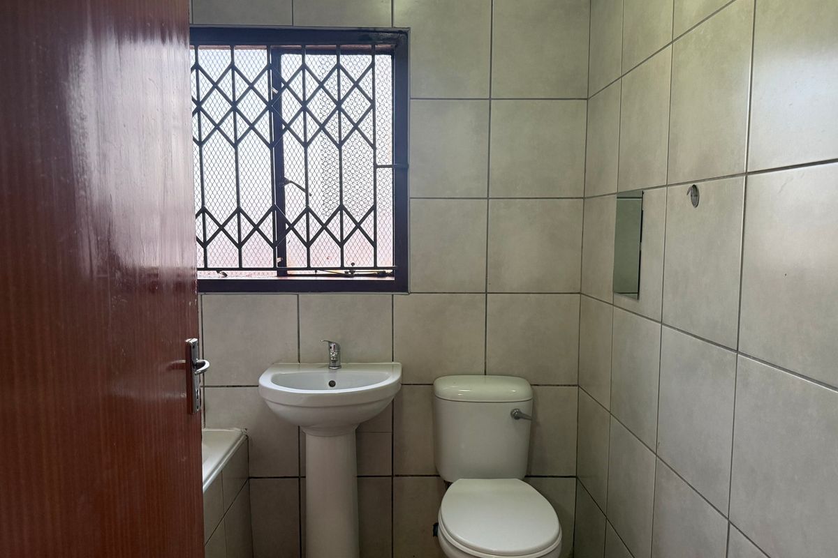 A functional bathroom featuring a pedestal basin, toilet, and bathtub, finished with neutral wall tiling and a security-gated window.