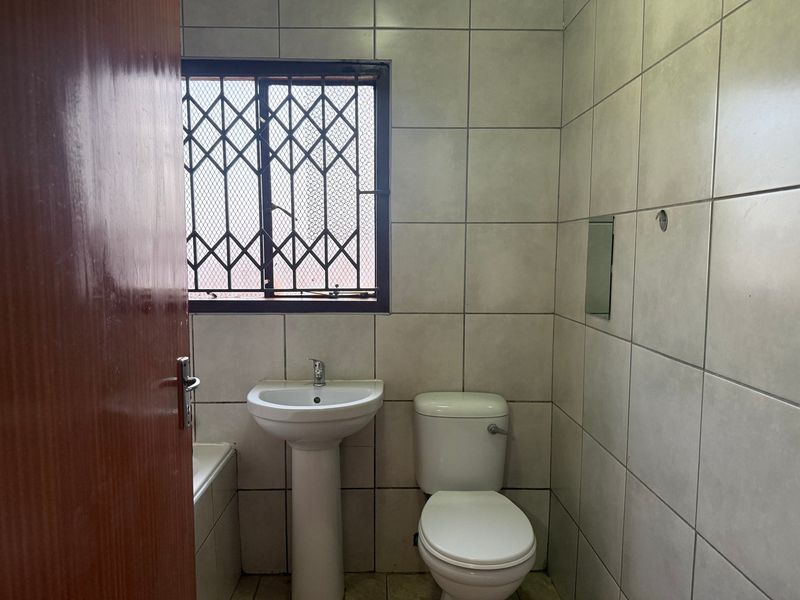A functional bathroom featuring a pedestal basin, toilet, and bathtub, finished with neutral wall tiling and a security-gated window.