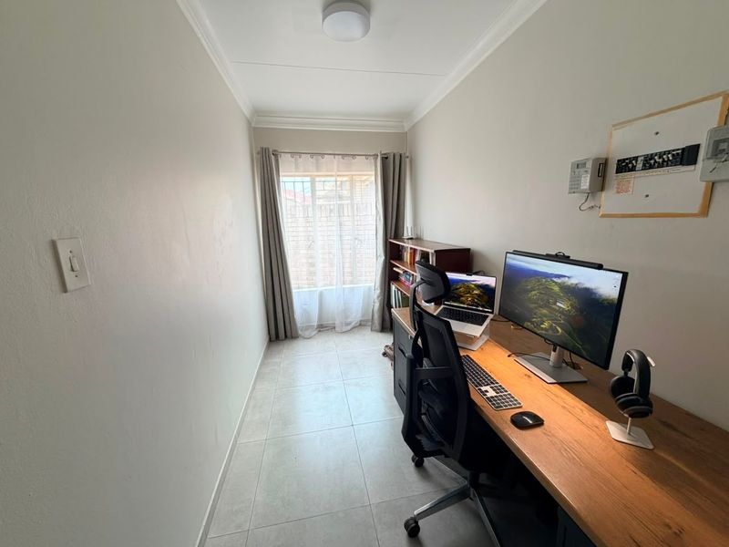 A functional home office space featuring tiled flooring, security bars on the window, and a wall-mounted electrical distribution board.