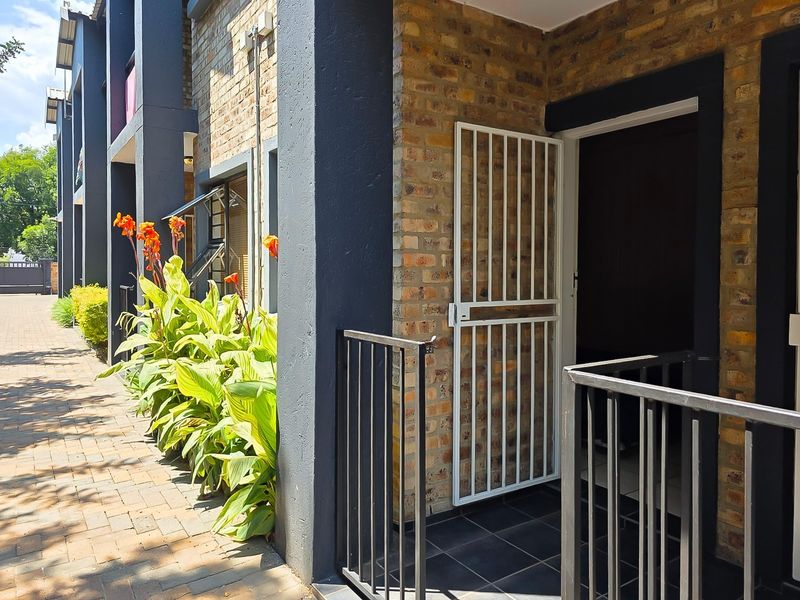 Front entrance of the apartment featuring a brick facade, secure metal gate, and a paved walkway with landscaped garden beds.