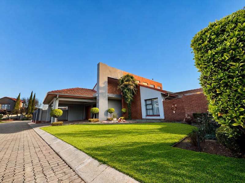 A well-maintained front exterior view of a house featuring a brick facade, tiled roof, paved driveway, and a neatly landscaped front lawn.