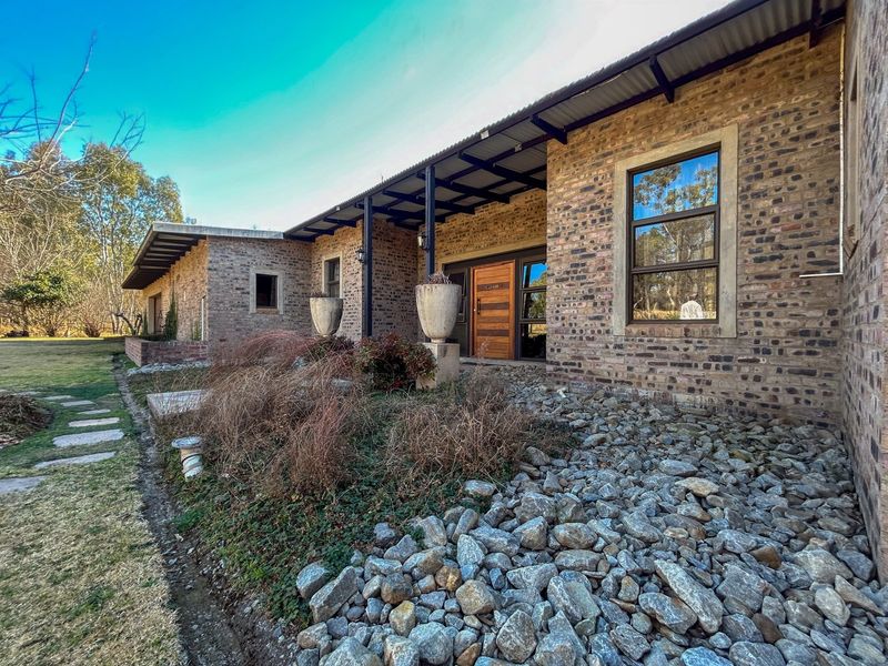 A charming single-story home featuring an exposed brick facade, a covered veranda, and a timber entrance door, set within a natural landscaped garden.