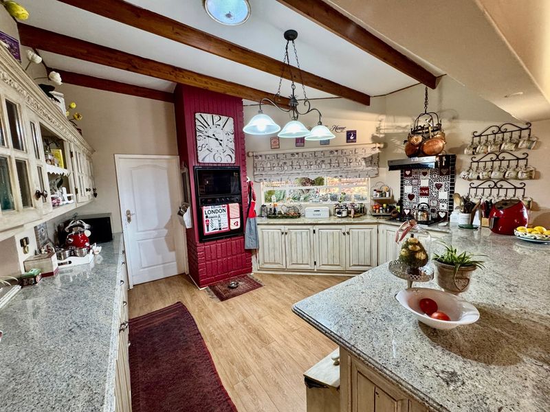 A rustic-style kitchen featuring granite countertops, a central island, exposed timber ceiling beams, and a built-in oven set within a red brick feature wall.