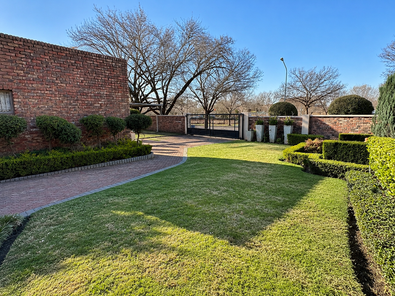 A well-maintained front garden featuring a paved driveway, manicured hedges, and a secure brick boundary wall with a metal entrance gate.