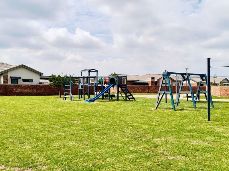 A spacious communal garden area featuring a well-maintained lawn and fixed playground equipment, enclosed by a secure brick boundary wall.