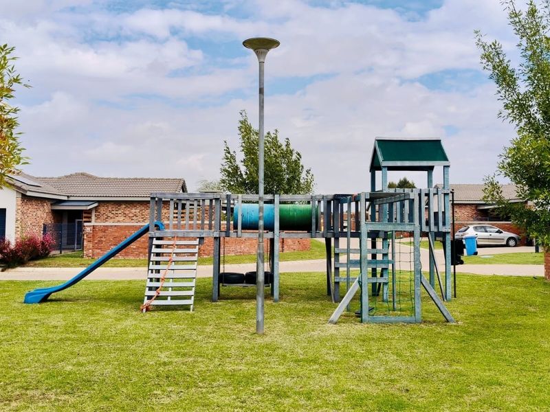 A communal garden area featuring a wooden playground structure, manicured lawn, and brick boundary walls within a residential estate.