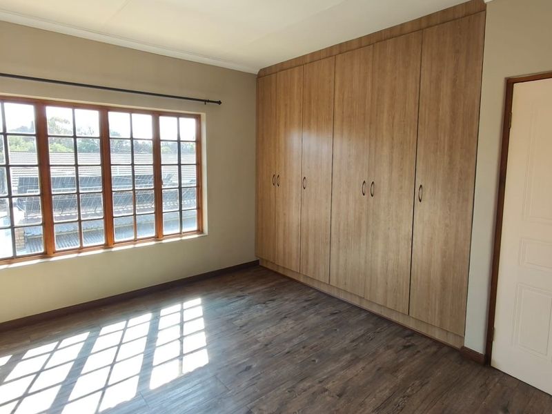 A bright, empty bedroom featuring laminate flooring, large wooden-framed windows, and extensive built-in cupboard storage.