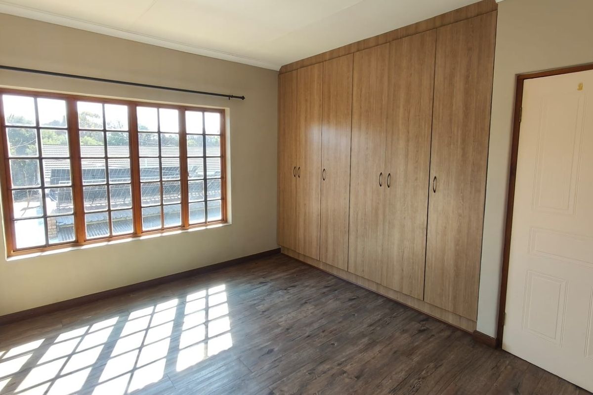 A bright, empty bedroom featuring laminate flooring, large wooden-framed windows, and extensive built-in cupboard storage.