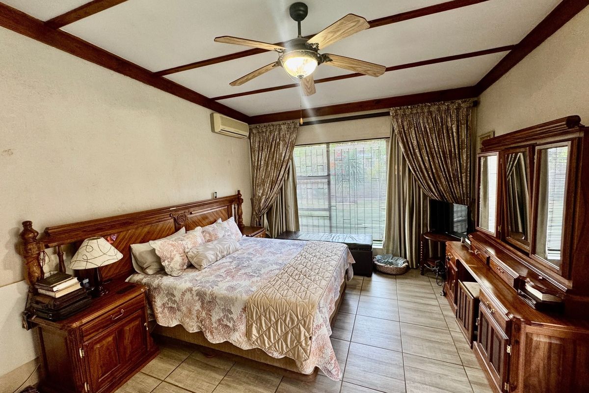 A spacious bedroom featuring tiled flooring, exposed timber ceiling beams, a ceiling fan, and a wall-mounted split-system air conditioner.