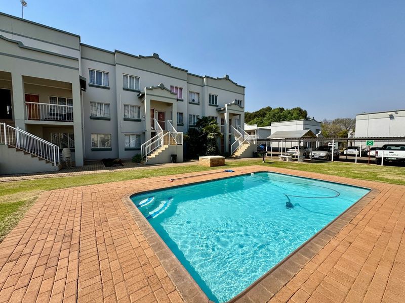 Communal swimming pool area featuring brick paving, well-maintained lawn, and access to apartment building staircases and covered parking bays.
