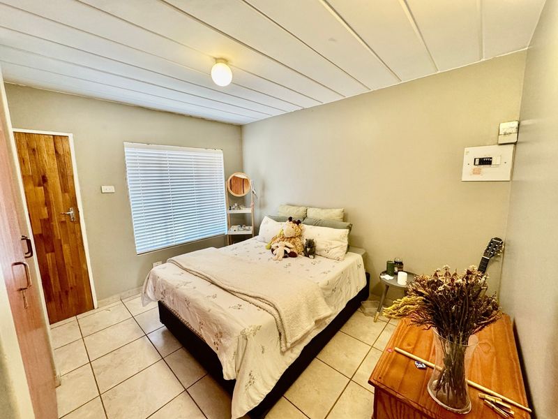 A functional bedroom featuring tiled flooring, timber door, and venetian blinds, with a neutral wall finish and panelled ceiling.