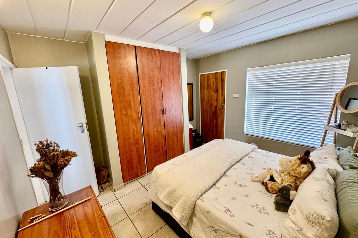 A functional bedroom featuring built-in wooden cupboards, tiled flooring, and a panelled ceiling with natural light provided by a window with venetian blinds.