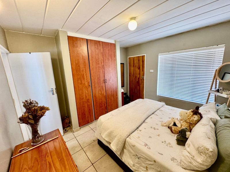 A functional bedroom featuring built-in wooden cupboards, tiled flooring, and a panelled ceiling with natural light provided by a window with venetian blinds.