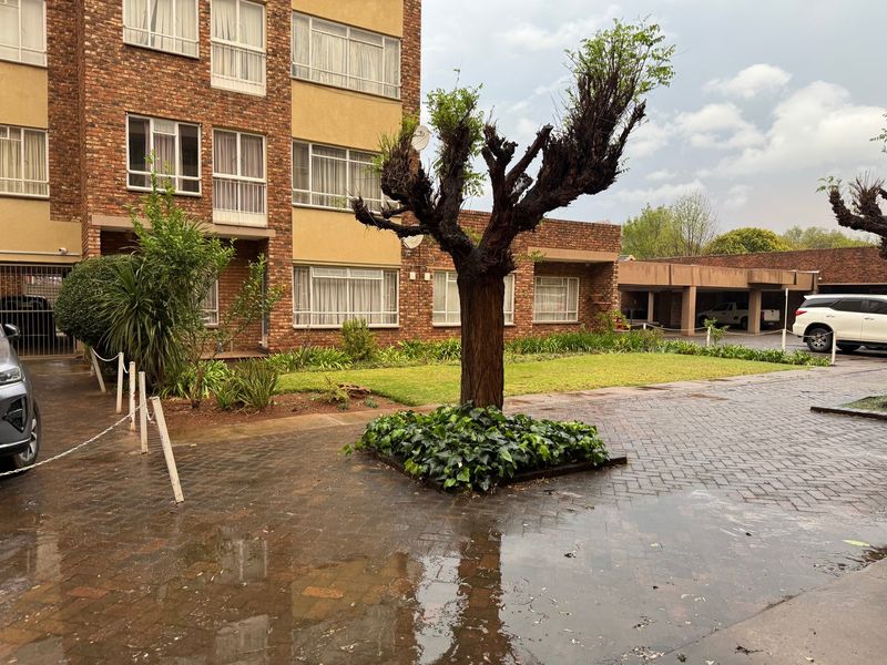 Exterior view of an apartment complex featuring a brick facade, paved driveway, landscaped garden area, and covered parking facilities.