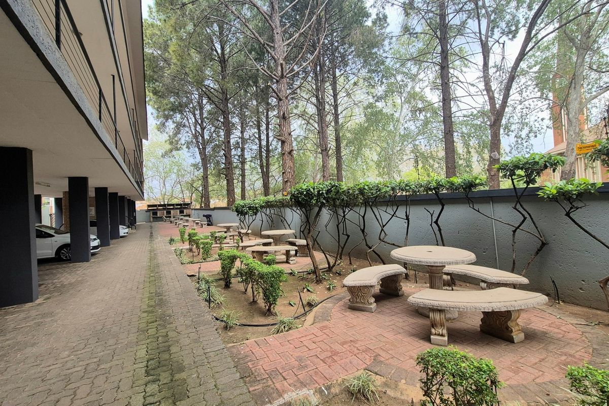 A communal outdoor area featuring brick-paved pathways, landscaped garden beds, and a boundary wall, situated alongside the apartment building's parking area.