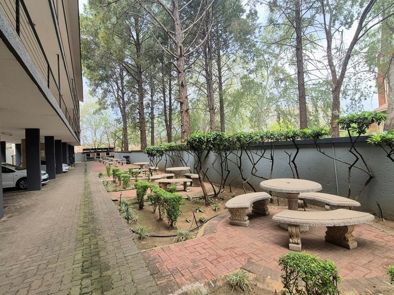 A communal outdoor area featuring brick-paved pathways, landscaped garden beds, and a boundary wall, situated alongside the apartment building's parking area.