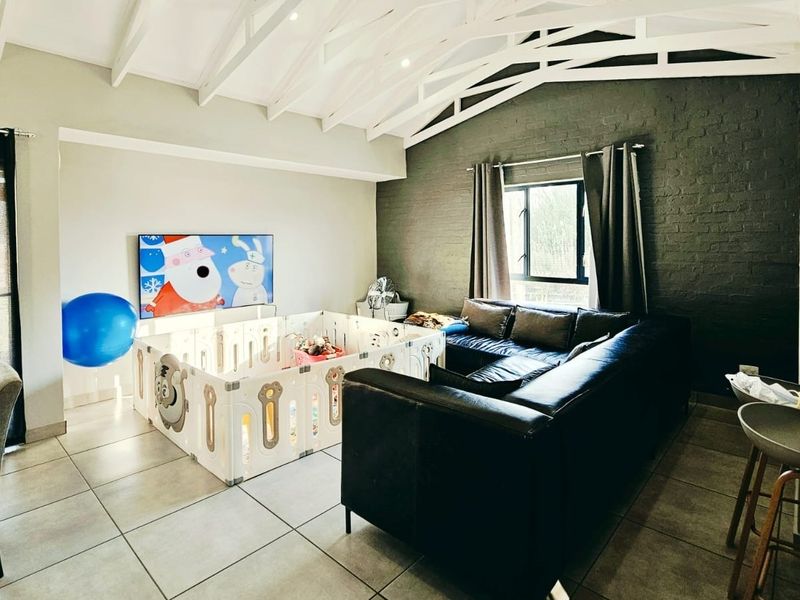 Spacious open-plan living area featuring high vaulted ceilings with exposed trusses, tiled flooring, and a striking dark exposed brick feature wall.