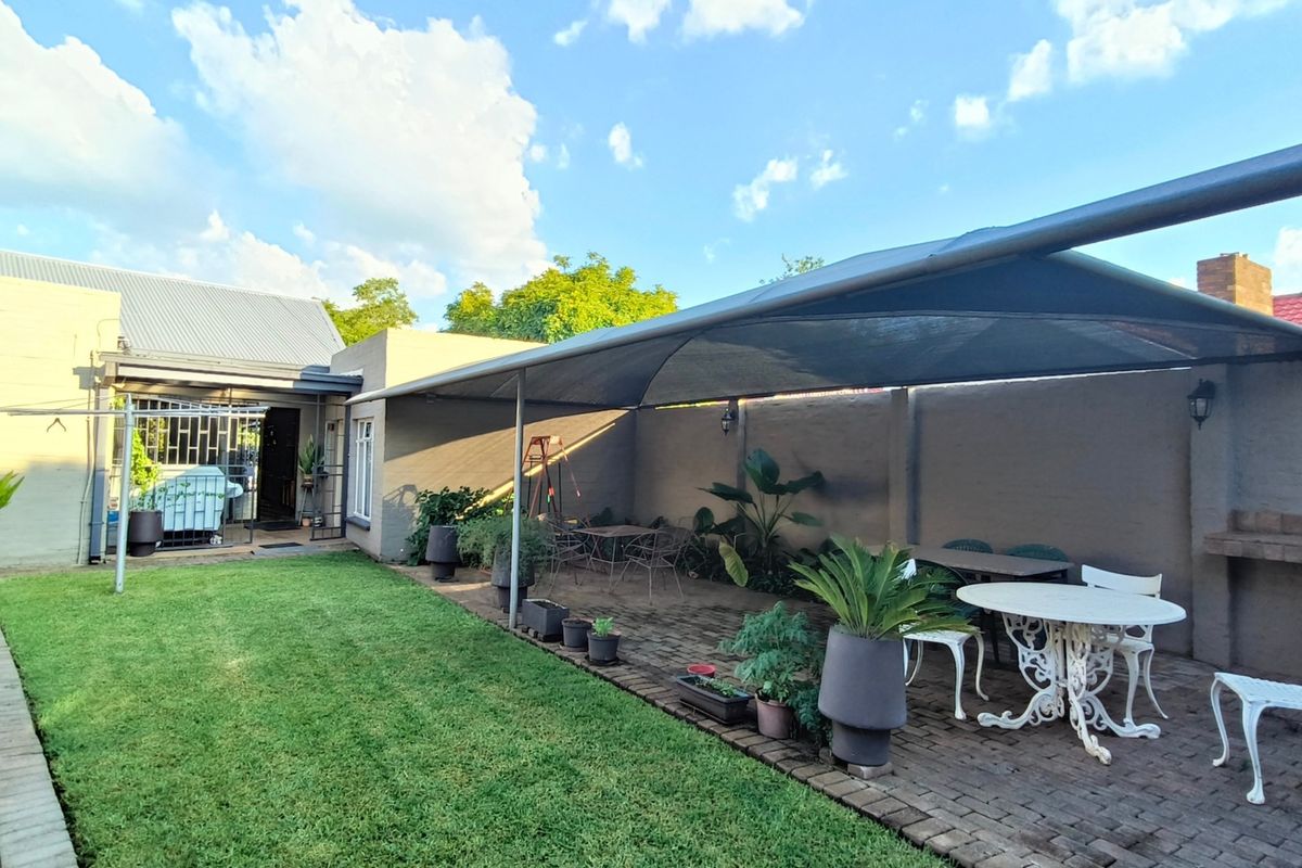 A spacious rear garden featuring a brick-paved patio area covered by a shade sail, complete with a built-in braai and well-maintained lawn.