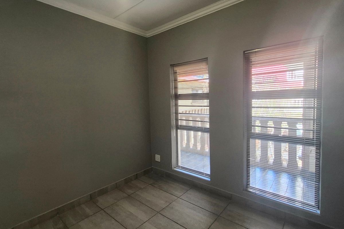 A clean, empty bedroom featuring neutral grey walls, tiled flooring, and two windows fitted with venetian blinds.