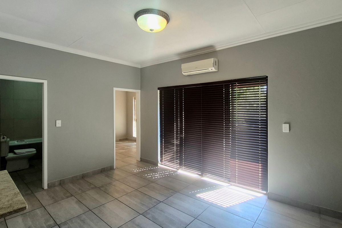 A bright, open-plan living area featuring neutral grey walls, tiled flooring, a wall-mounted air conditioning unit, and large windows with fitted venetian blinds.
