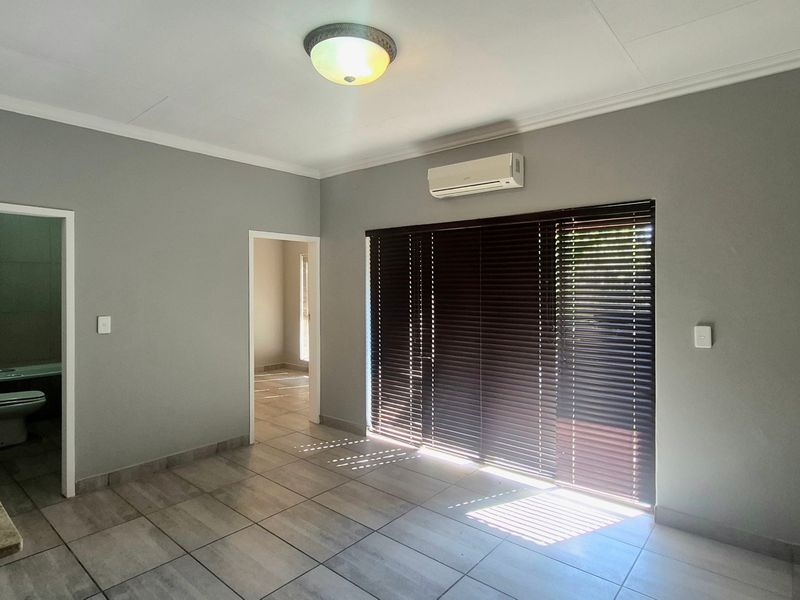 A bright, open-plan living area featuring neutral grey walls, tiled flooring, a wall-mounted air conditioning unit, and large windows with fitted venetian blinds.