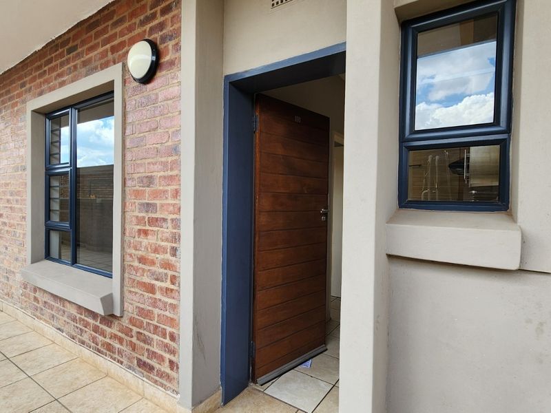 Front entrance of the apartment featuring a solid timber door, brick facade detailing, and tiled patio area with modern aluminium window frames.
