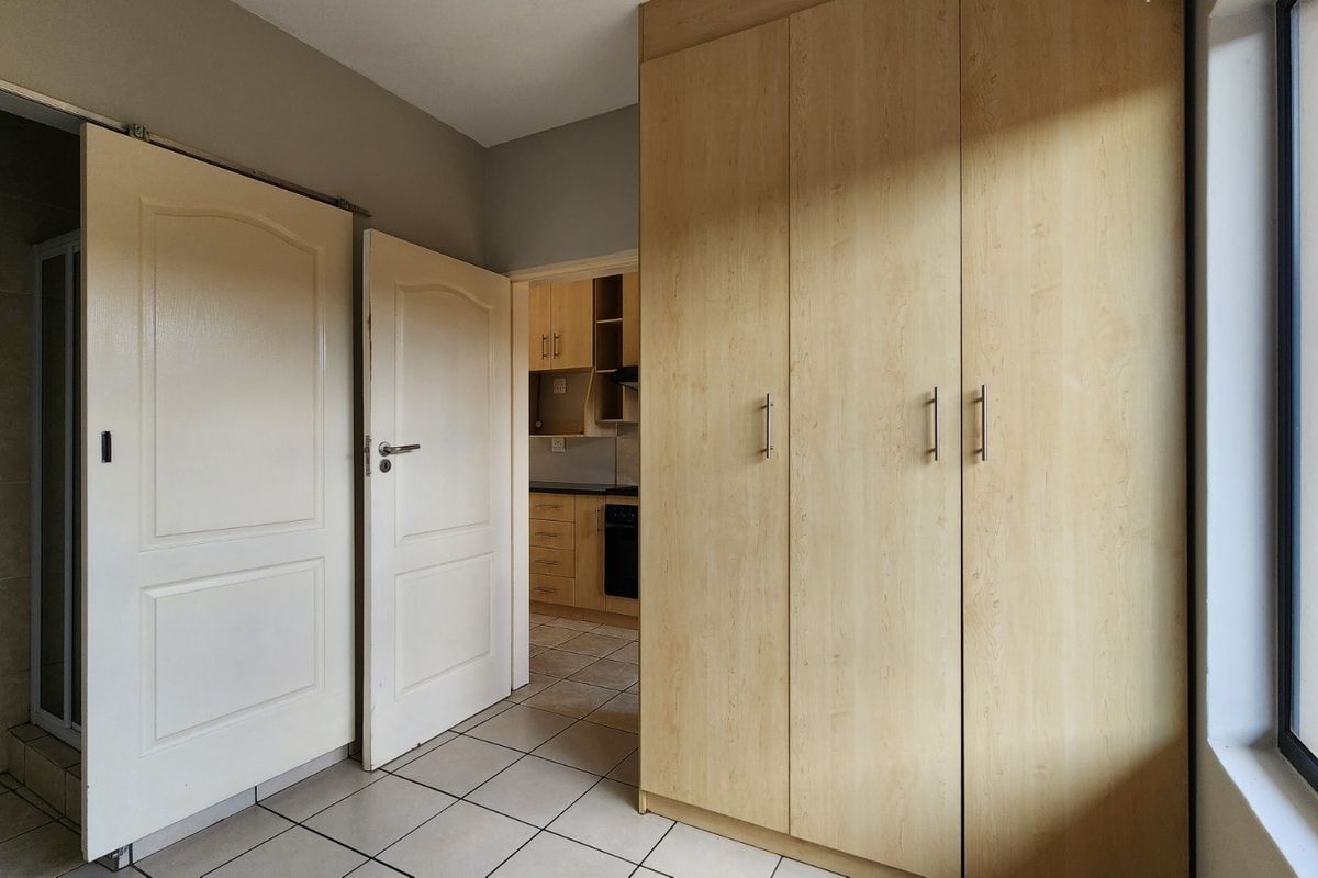 A bright bedroom featuring built-in wooden cupboards, tiled flooring, and direct access to an adjacent kitchen area.