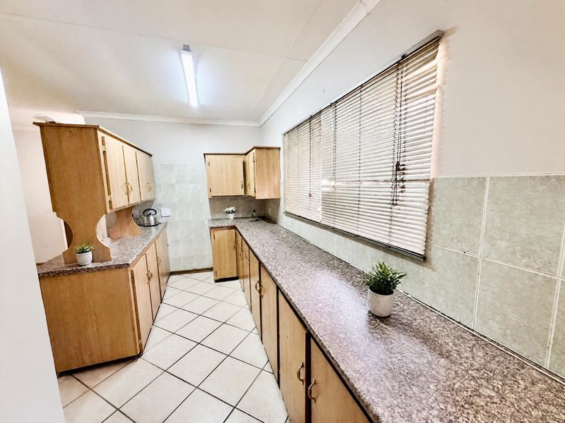 A functional kitchen featuring extensive laminate countertops, built-in wooden cabinetry, tiled splashbacks, and ceramic floor tiles with large windows.