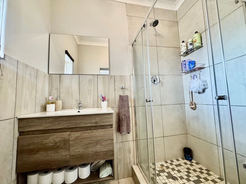 A modern bathroom featuring a wood-finish vanity cabinet, a wall-mounted mirror, and a glass-enclosed shower with neutral-toned wall tiles.