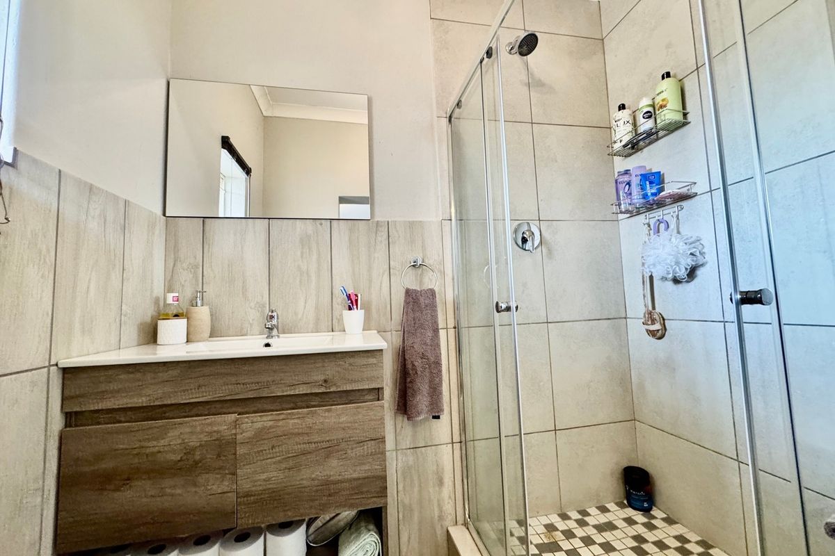 A modern bathroom featuring a wood-finish vanity cabinet, a wall-mounted mirror, and a glass-enclosed shower with neutral-toned wall tiles.