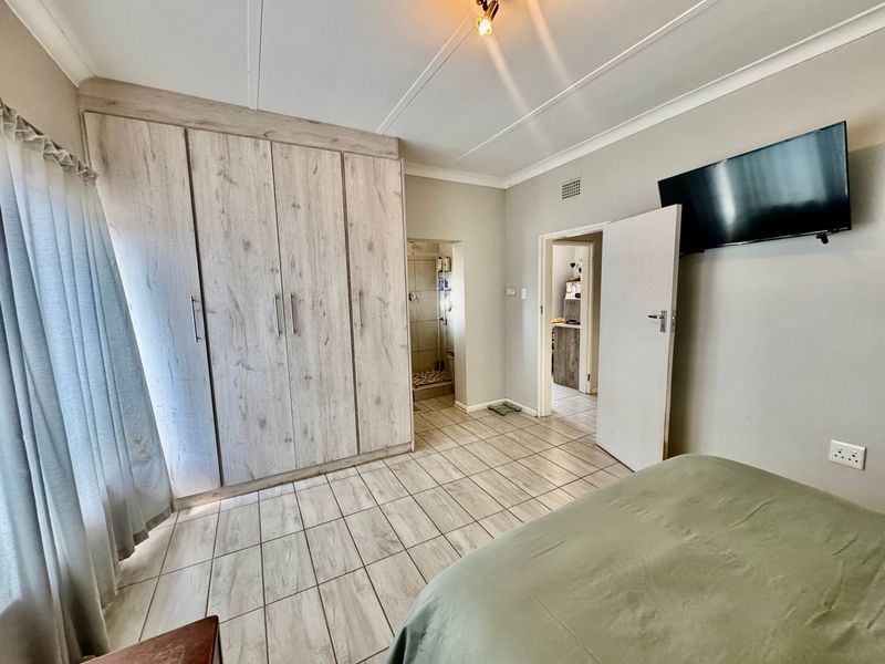A well-maintained bedroom featuring light wood-look built-in cupboards, neutral tiled flooring, and direct access to an en-suite bathroom.