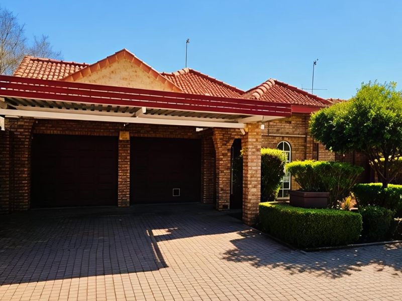 Front view of a townhouse featuring a double garage, paved driveway, carport, and well-maintained brick facade with established garden landscaping.