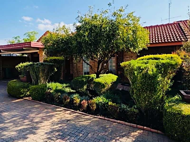 Front elevation of a townhouse featuring a brick facade, tiled roof, paved driveway, and a covered carport area with established garden landscaping.