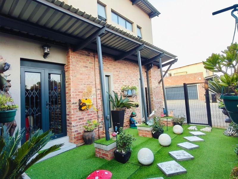 A well-maintained covered patio area featuring exposed brickwork, steel structural supports, and a low-maintenance artificial lawn with decorative stone pavers.