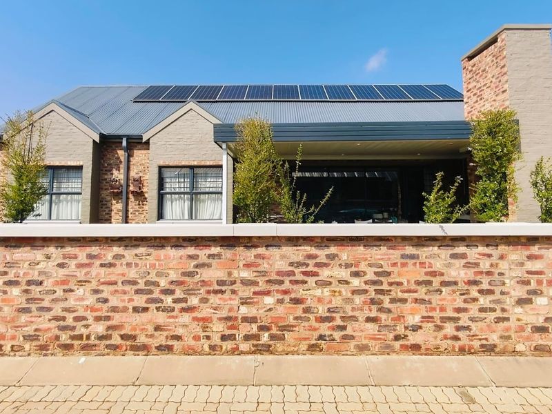 Front view of a modern brick house featuring a covered patio, solar panels on a corrugated metal roof, and a solid brick boundary wall.